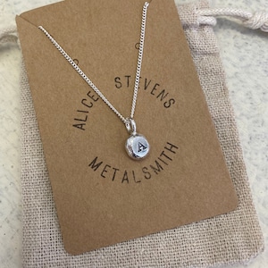 May include: A silver necklace with a small, round pendant featuring the letter "A" engraved on it. The necklace is presented in a brown paper bag with the text "Alice Stevens Metalsmith" printed on it.
