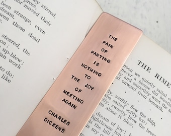 May include: A copper bookmark with a quote about the pain of parting and the joy of meeting again. The quote reads "The pain of parting is nothing to the joy of meeting again". The bookmark also features the name "Charles Dickens" at the bottom.