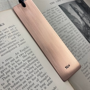 May include: A rose gold bookmark with a black leather tassel. The bookmark has the name "BEN" engraved on it. The bookmark is lying on top of an open book.