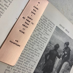 May include: A copper bookmark with the text "If I loved you less, I might be able to talk about it more." and "Mr. Knightley" on the front. The bookmark is lying on top of an open book.