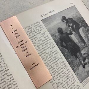 May include: A copper bookmark with an engraved quote: "If I loved you less, I might be able to talk about it more." The quote is attributed to Mr. Knightley. The bookmark is lying on a page of a book titled "Adam Bede".