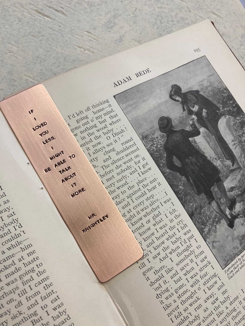 May include: A copper bookmark with an engraved quote: "If I loved you less, I might be able to talk about it more." The quote is attributed to Mr. Knightley. The bookmark is lying on a page of a book titled "Adam Bede".