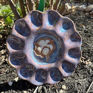 May include: A handcrafted ceramic dish with a scalloped edge and a cotton blossom design in the center. The dish features a blend of blue, purple, and brown hues, creating a unique, iridescent finish. This decorative piece is perfect for serving or display.