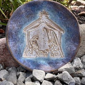 May include: A round ceramic plate featuring a glazed finish in shades of blue and purple. The center displays an embossed nativity scene with a stable, a star, and figures, rendered in a light tan color with subtle speckles across the surface.