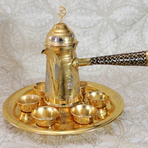 May include: A gold-plated Arabic coffee pot with a black handle and six small gold cups on a gold tray.