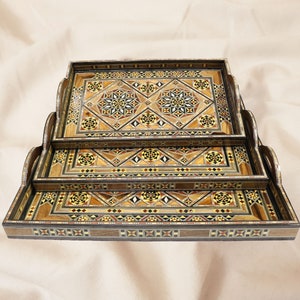 Puede incluir: Tres bandejas de madera con intrincados patrones geométricos en tonos marrones, beige y negros. Las bandejas están apiladas una encima de la otra, con la bandeja más grande en la parte inferior y la bandeja más pequeña en la parte superior.