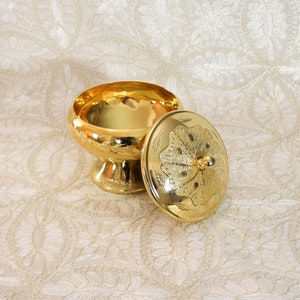 Könnte beinhalten: Ein goldener Metallbehälter mit Deckel. Der Behälter hat ein verziertes Design und einen runden Boden. Der Deckel hat ein Blumenmuster.