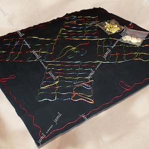 Puede incluir: Un tablero de fieltro negro con bordados coloridos en forma de pájaro. El tablero está rodeado por un borde cosido rojo. Hay dos pequeñas bolsas de suministros de artesanía, una con cuentas doradas y la otra con cuentas blancas.