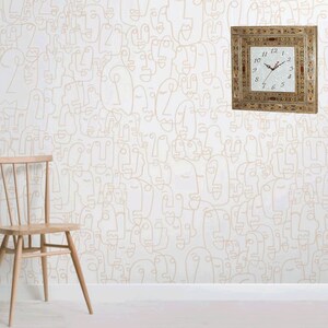 May include: A white wall with a beige patterned wallpaper featuring abstract faces. A square wooden clock with Roman numerals and a white face is mounted on the wall. A wooden chair is in the foreground.