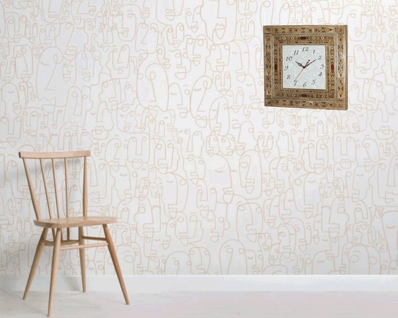 May include: A white wall with a beige patterned wallpaper featuring abstract faces. A square wooden clock with Roman numerals and a white face is mounted on the wall. A wooden chair is in the foreground.