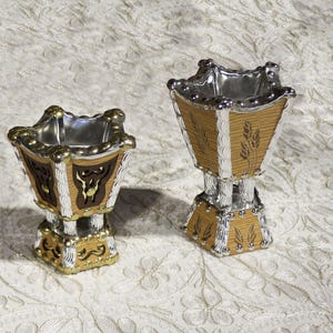 May include: Two decorative incense burners. The burners have a square shape with a silver interior and a gold and silver exterior. The exterior features a wood-like pattern with decorative accents. One burner has a bull design, and the other has a wheat design.