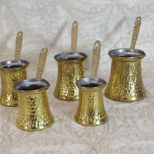 May include: Set of five gold-colored hammered metal coffee pots with long handles. The pots are of varying sizes and are arranged on a white fabric surface.
