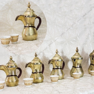 May include: A set of six gold-colored metal coffee pots with ornate designs and brown leather handles. Each pot is paired with a small white cup with gold accents. The cups and pots are arranged on a white surface with a floral pattern.