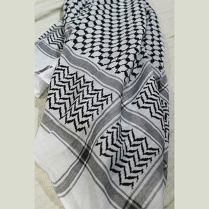 Könnte beinhalten: Ein weiß-schwarzes Kopftuch mit traditionellem geometrischem Muster. Das Tuch ist gefaltet und über eine Oberfläche gelegt.