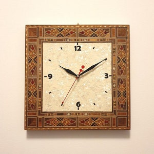 May include: A square clock with a white face and black hands. The clock is surrounded by a decorative frame made of inlaid wood with geometric patterns in shades of brown, red, and gold.