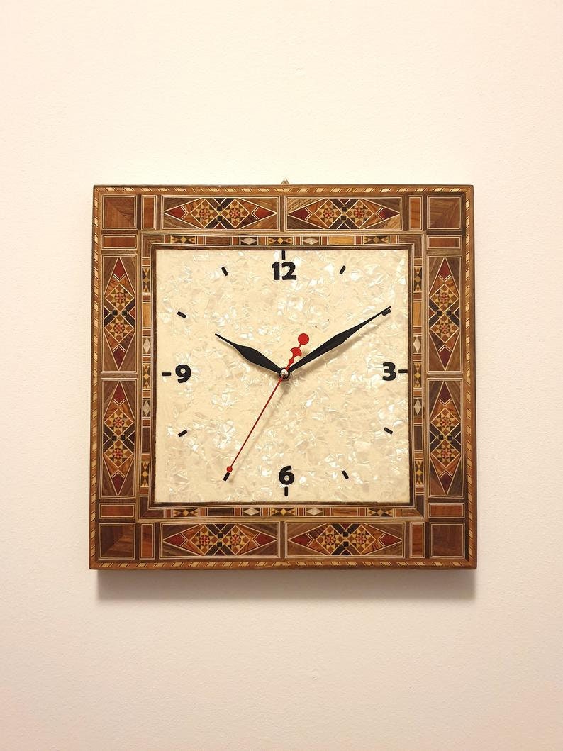 May include: A square clock with a white face and black hands. The clock is surrounded by a decorative frame made of inlaid wood with geometric patterns in shades of brown, red, and gold.