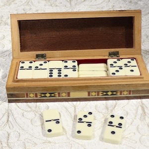 May include: A wooden domino set in a box with a hinged lid. The dominoes are cream-coloured with black dots. The box has inlaid patterns and a red felt lining. Three dominoes are placed outside the box.
