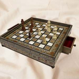 May include: A wooden chessboard with inlaid mother-of-pearl squares. The board has a drawer for storing the pieces. The pieces are arranged on the board in a game in progress.
