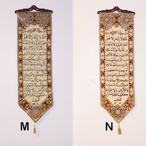 May include: Two decorative wall hangings with Arabic calligraphy. Each rectangular tapestry features a dark brown wooden top with a decorative pattern, a central text panel, and a tassel. The text is in black against a cream background, with ornate borders.