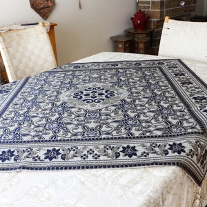 May include: A white tablecloth with a blue and gray floral pattern. The tablecloth has a square shape and a fringed edge.