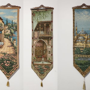 May include: Three vertical woven fabric wall hangings with pointed bottoms and gold tassels. The left depicts a coastal path with flowers, the center shows an arched courtyard with a fountain, and the right features a mosque with minarets.