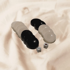 Puede incluir: Piezas de juego de backgammon, que incluyen fichas circulares negras y blancas y dos dados. Las fichas están dispuestas en dos grupos, con los dados colocados cerca. Las piezas del juego están sobre una tela color crema.