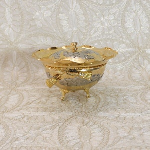 Könnte beinhalten: Eine dekorative Schale aus Gold und Silber mit Deckel. Die Schale hat einen gewellten Rand und steht auf drei Füßen. Der Deckel hat ein Blumenmuster.