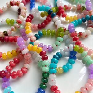 May include: A selection of multi-coloured beaded bracelets, featuring a spectrum of hues such as red, yellow, green, blue, and purple. The beads are threaded on red string, creating a vibrant display. The bracelets are arranged on a white surface.