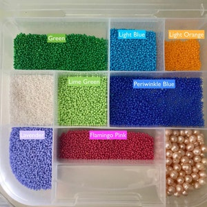 May include: A clear plastic storage box with compartments filled with various colours of small round beads. The compartments are labelled with the colour of the beads: Green, Light Blue, Light Orange, Lime Green, Periwinkle Blue, Lavender, Flamingo Pink. The last compartment contains larger, round, pearl-like beads.