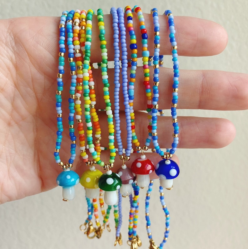 May include: A set of colourful beaded necklaces with mushroom charms. The necklaces are made with small, colourful beads and feature a variety of colours, including blue, green, yellow, orange, and red. Each necklace has a gold clasp and a small mushroom charm.