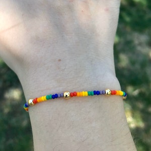 May include: A rainbow beaded bracelet with gold beads. The bracelet is on a person's wrist.