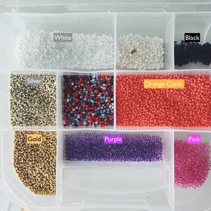 May include: A clear plastic storage box with compartments filled with different coloured beads. The compartments are labelled "White", "Silver", "Gold", "Orange Coral", "Purple", "Pink", and "Black".