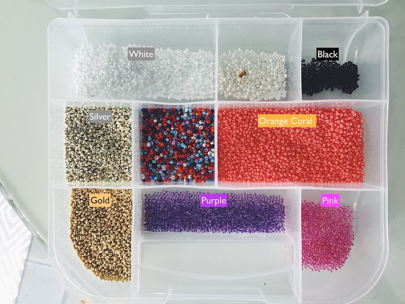 May include: A clear plastic storage box with compartments filled with different coloured beads. The compartments are labelled "White", "Silver", "Gold", "Orange Coral", "Purple", "Pink", and "Black".