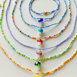May include: Seven colourful beaded necklaces with mushroom charms. The necklaces are layered in a rainbow pattern, with the shortest necklace at the bottom and the longest at the top. The mushroom charms are made of glass and have different colours and patterns.