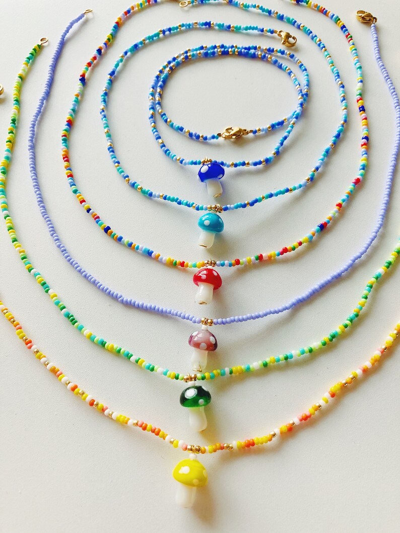 May include: Seven colourful beaded necklaces with mushroom charms. The necklaces are layered in a rainbow pattern, with the shortest necklace at the bottom and the longest at the top. The mushroom charms are made of glass and have different colours and patterns.