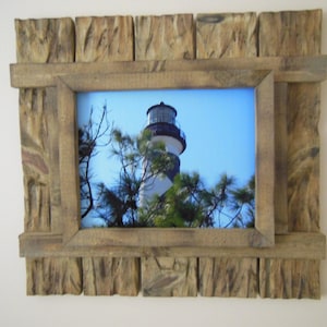 May include: A rustic wooden picture frame with a lighthouse photograph inside. The frame is made of reclaimed wood with a natural finish. The lighthouse is white with a black top and is surrounded by green trees.