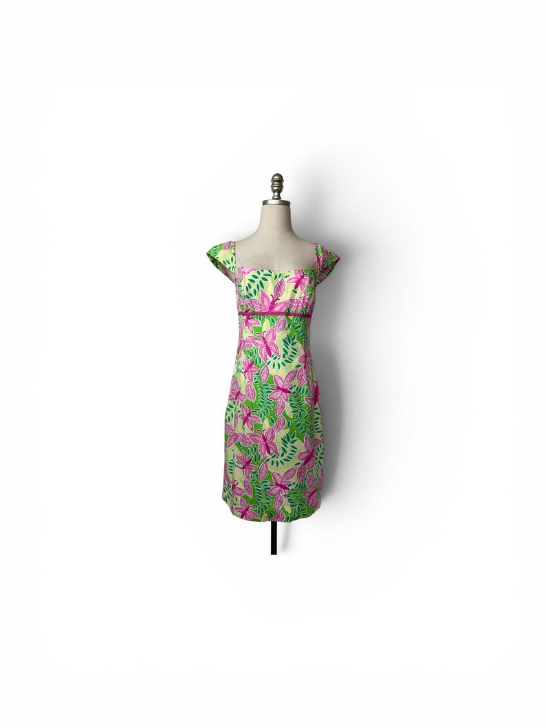 May include: A knee-length dress featuring a vibrant pattern of pink butterflies and green leaves on a light yellow background. The garment has short cap sleeves, a square neckline, and a solid dark pink horizontal band at the waistline.