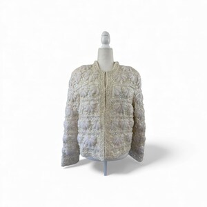 May include: A cream-colored jacket with floral embellishments and sparkling sequins. The jacket is displayed on a mannequin and features a round neckline and long sleeves. The jacket is fully covered in decorative elements.