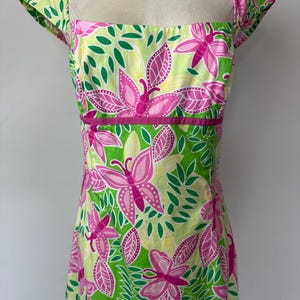 May include: A sleeveless dress featuring a square neckline and cap sleeves, decorated with a vibrant pattern of pink butterflies and green leaves on a light yellow background, accented by a solid pink horizontal band at the waistline.