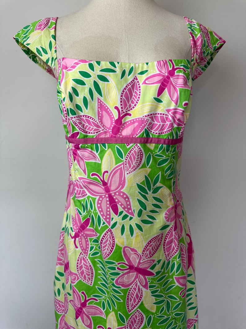 May include: A sleeveless dress featuring a square neckline and cap sleeves, decorated with a vibrant pattern of pink butterflies and green leaves on a light yellow background, accented by a solid pink horizontal band at the waistline.