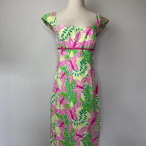 May include: A knee-length dress featuring a vibrant pattern of pink butterflies and green leaves on a pale yellow background. The garment has a square neckline, short cap sleeves, and a fitted bodice with a horizontal pink band at the waist.