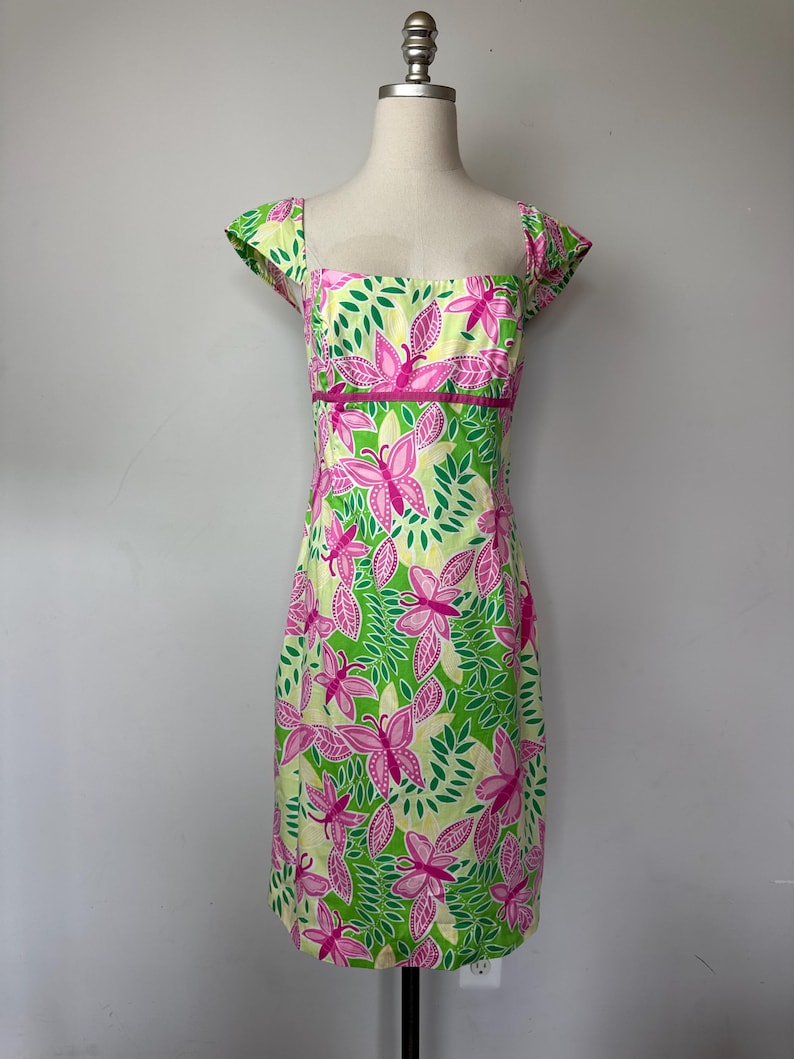 May include: A knee-length dress featuring a vibrant pattern of pink butterflies and green leaves on a pale yellow background. The garment has a square neckline, short cap sleeves, and a fitted bodice with a horizontal pink band at the waist.