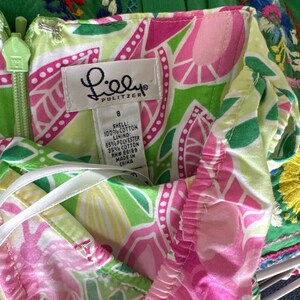 May include: A Lilly Pulitzer dress featuring a vibrant floral pattern in shades of lime green, hot pink, and white. The garment includes a size 8 tag, a light green zipper, and a label indicating a shell of 100% cotton and a lining of 65% polyester and 35% cotton.