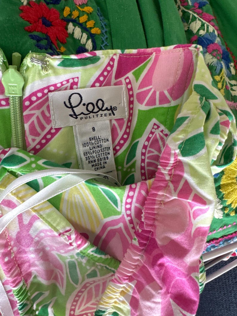 May include: A Lilly Pulitzer dress featuring a vibrant floral pattern in shades of lime green, hot pink, and white. The garment includes a size 8 tag, a light green zipper, and a label indicating a shell of 100% cotton and a lining of 65% polyester and 35% cotton.