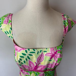 May include: A sleeveless dress featuring a vibrant pattern of pink butterflies, green leaves, and yellow accents on a light green background. The garment has a square neckline, cap sleeves, and a horizontal pink band detail at the waistline.