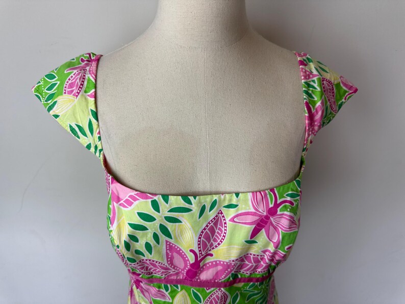 May include: A sleeveless dress featuring a vibrant pattern of pink butterflies, green leaves, and yellow accents on a light green background. The garment has a square neckline, cap sleeves, and a horizontal pink band detail at the waistline.