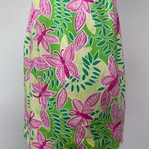 May include: A sleeveless dress featuring a vibrant pattern of pink butterflies, green leaves, and pale yellow accents against a bright lime green background. The garment has a straight silhouette and a repeating botanical and insect print design.