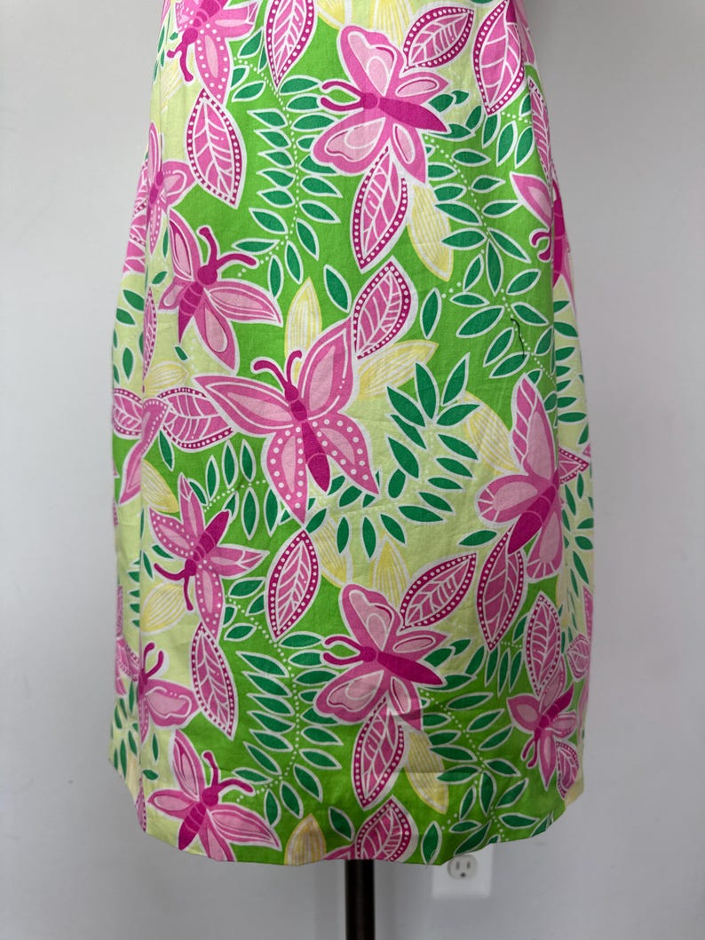 May include: A sleeveless dress featuring a vibrant pattern of pink butterflies, green leaves, and pale yellow accents against a bright lime green background. The garment has a straight silhouette and a repeating botanical and insect print design.