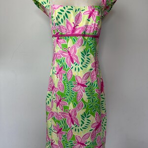 May include: A knee-length dress featuring a vibrant pattern of pink butterflies and green leaves on a pale yellow background. The garment has a square neckline, short cap sleeves, and a solid pink horizontal band accenting the waistline.