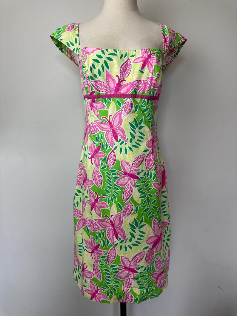 May include: A knee-length dress featuring a vibrant pattern of pink butterflies and green leaves on a pale yellow background. The garment has a square neckline, short cap sleeves, and a solid pink horizontal band accenting the waistline.